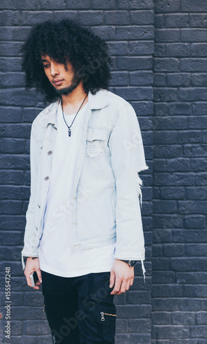 Young black man with afro hair posing on graphite background. Outside. Dressed in a jeans jacket, black trousers. Fashion. Street style.