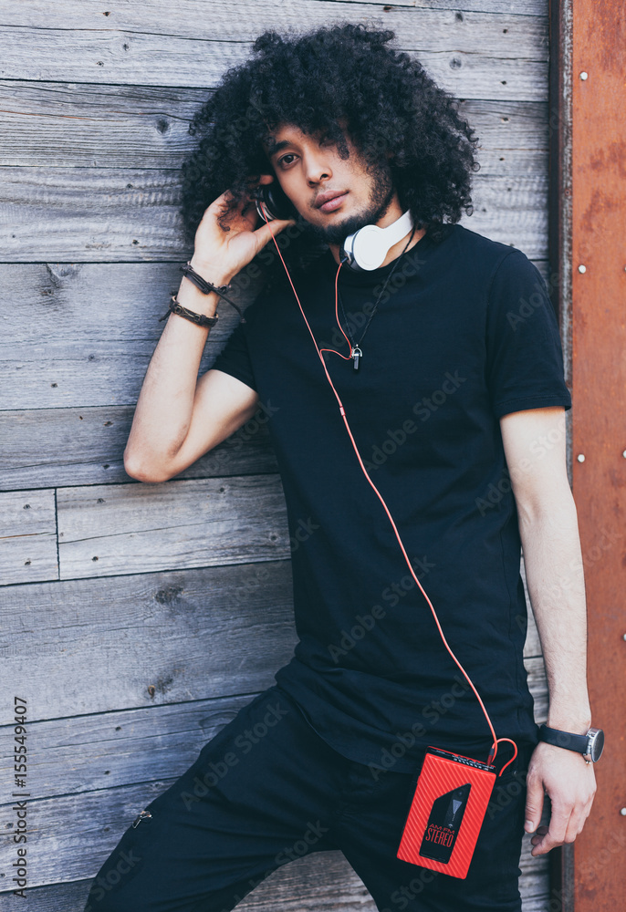 Close up portrait African American man with an afro hairstyle listens to music as DJ, resting. Headphones and vintage cassette player. Wearing a black outfit and high white sneakers. Rockstar. Reggae. Stock Close up portrait African American man with an afro hairstyle listens to music as DJ, resting. Headphones and vintage cassette player. Wearing a black outfit and high white sneakers. Rockstar. Reggae. Stock