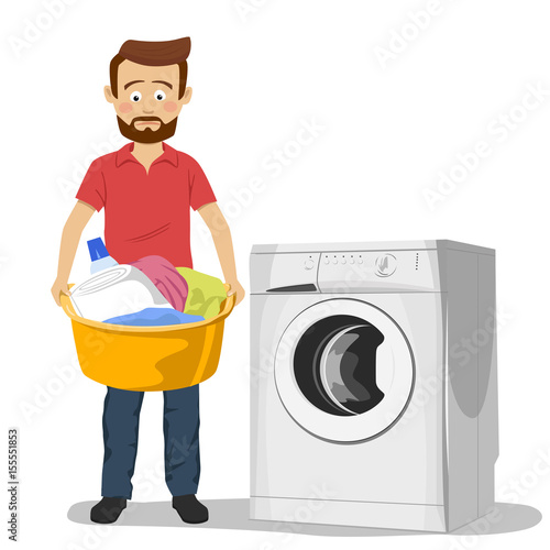 Unhappy young man standing next to washing machine with basin filled with dirty clothes