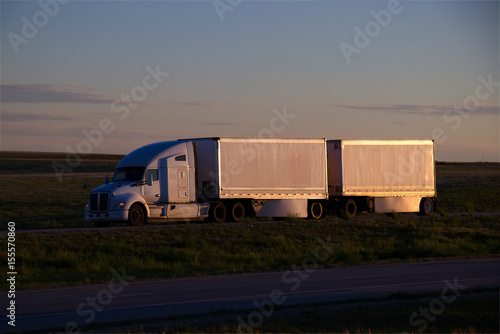 A white Kenworth sleeper truck pulls a set of white pup doubles down a rural US highway during sunset hours. All visible trademarks and markings have been removed. 