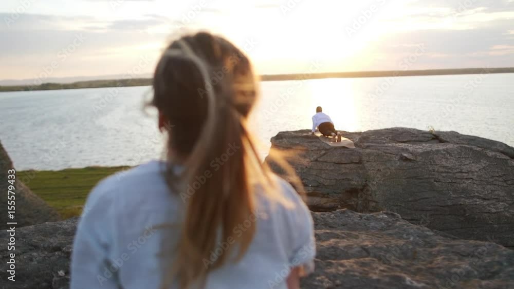 Beautiful young sporty man and woman doing ashtanga yoga on the rock outdoor in front of the lake sunrise. Freedom, health and yoga concept. Handheld camera Beautiful young sporty man and woman doing ashtanga yoga on the rock outdoor in front of the lake sunrise. Freedom, health and yoga concept. Handheld camera