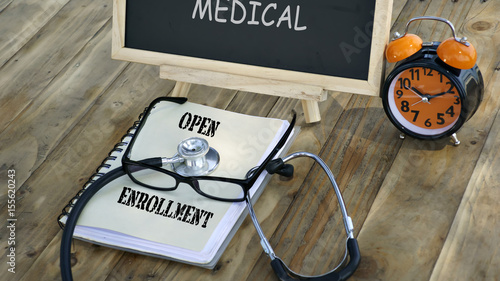 notebook with the words "OPEN ENROLLMENT" and stethoscope, glasses, chalk board, alarm clock on a wooden table. medical and healthcare concept.