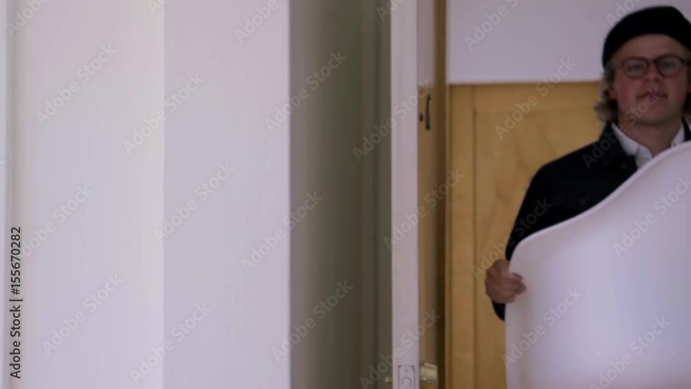 Young Couple Enter Their New Apartment, Carrying Furniture And A Plant, Set It Down Young Couple Enter Their New Apartment, Carrying Furniture And A Plant, Set It Down