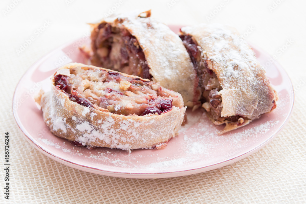Apple strudel with icing sugar, cherries and raisins on rosa plate Apple strudel with icing sugar, cherries and raisins on rosa plate