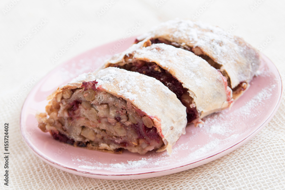 Apple strudel with icing sugar, cherries and raisins on rosa plate