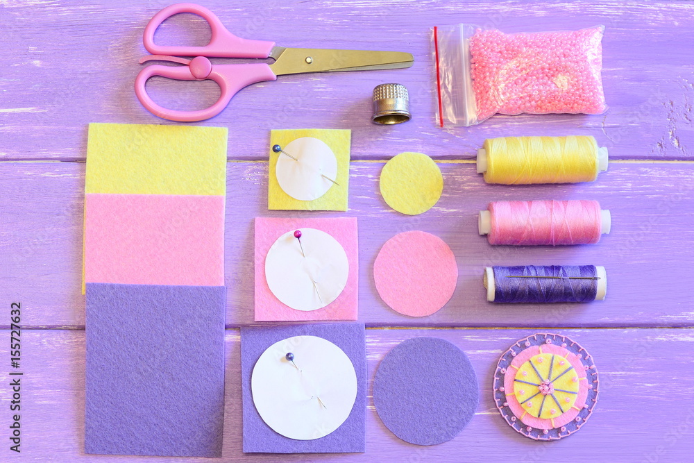 Obraz premium Cute flower made of felt and beads, colored felt circles and sheets, scissors, paper templates, thread spools, needle, pink beads on a wooden table. Sewing instruction. Teach children to sew. Top view