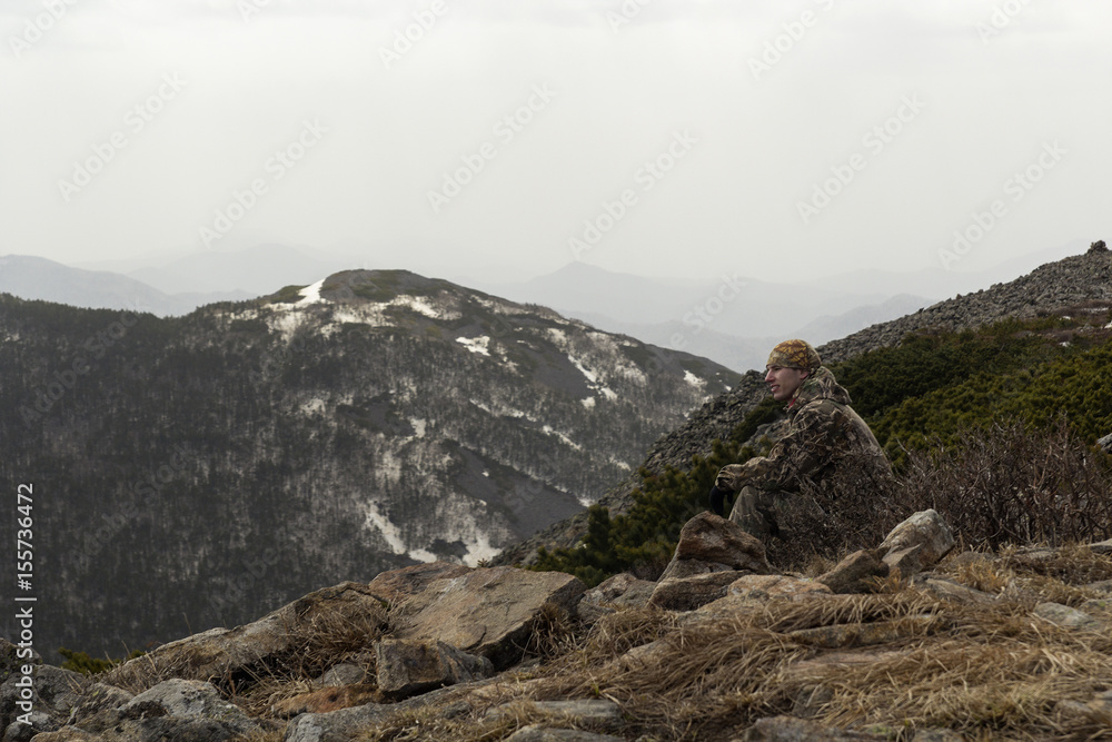 A man in camp clothes sits on a rock atop a mountain and admires the mountain scenery ahead