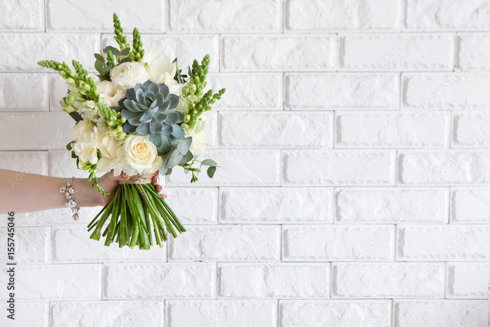 Fototapeta premium Female hand gives bunch with roses and succulent on white brick background. Gift for mother or woman, florist work, wedding decor, beautiful bouquet sale concept