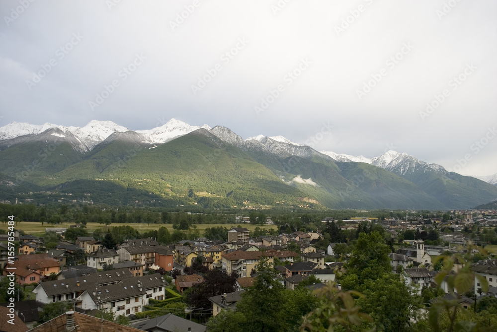 Landscape of mountains of a strong spring green, with snow-covered peaks after a strong thunderstorm of mountain and some rays of sunshine, in the foreground valley and houses , alps, italy