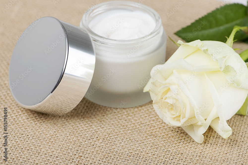 Close-up of a jar of cream for the skin and a white rose on brown rustic fabric. Horizontal shoot. Close-up of a jar of cream for the skin and a white rose on brown rustic fabric. Horizontal shoot.