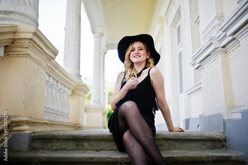 Blonde woman on black dress  necklaces and hat against vintage house.