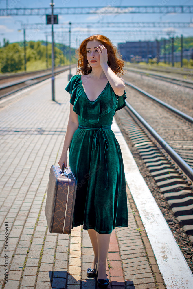 Gorgeous redhead woman in green velvet dress in vintage style with a suitcase on the platform
