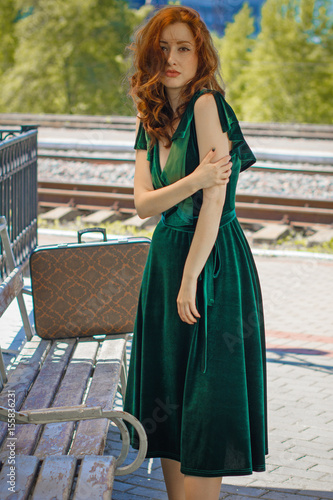 Gorgeous redhead woman in green velvet dress in vintage style with a suitcase on the platform