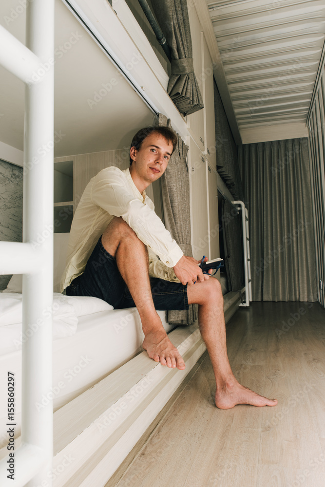 Man sitting in a stylish hostel bedroom. Young man reading book in hotel room with two-levels beds. Evening, grey curtain and wall Man sitting in a stylish hostel bedroom. Young man reading book in hotel room with two-levels beds. Evening, grey curtain and wall