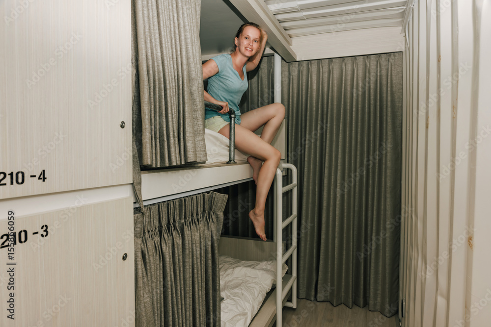 Girl sitting in a stylish hostel bedroom. Young woman sitting on second level bed in hotel room with two-levels beds. Evening, grey curtain and wall Girl sitting in a stylish hostel bedroom. Young woman sitting on second level bed in hotel room with two-levels beds. Evening, grey curtain and wall
