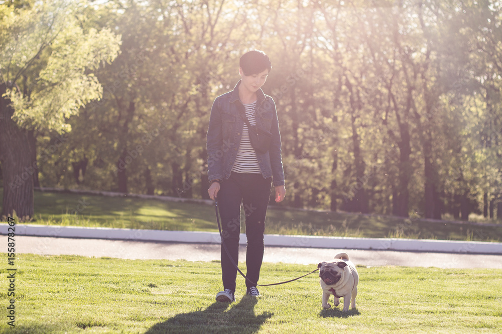 Young woman having good time with pug on the green grass, pretty girl with dog play in the park during sunset or sunrise Young woman having good time with pug on the green grass, pretty girl with dog play in the park during sunset or sunrise