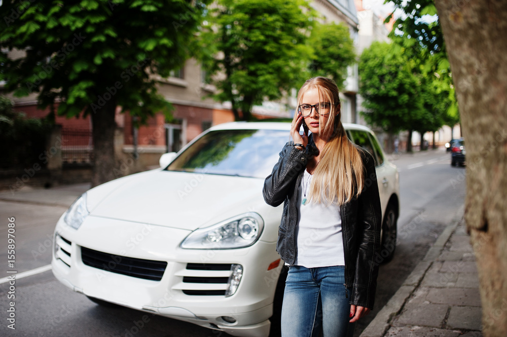 Stylish blonde woman wear at jeans, glasses and leather jacket with mobile phone, against luxury car. Fashion urban model portrait.