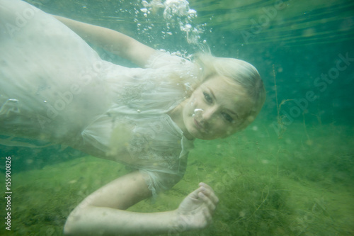 A floating woman. Underwater portrait. Girl in white dress swimming in the lake. Green marine plants and water