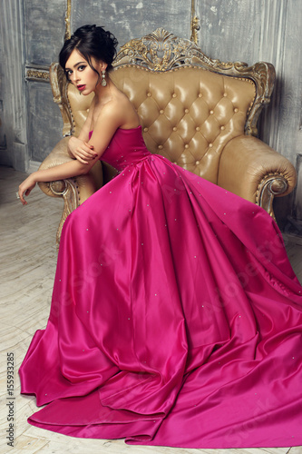 Elegant srunning woman sitting in armchair and looking at you. Pretty lady with hairstyle wearing pink ball gown or evening dress.