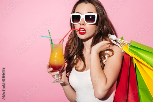 Girl in sunglasses holding colorful shopping bags and drinking cocktail