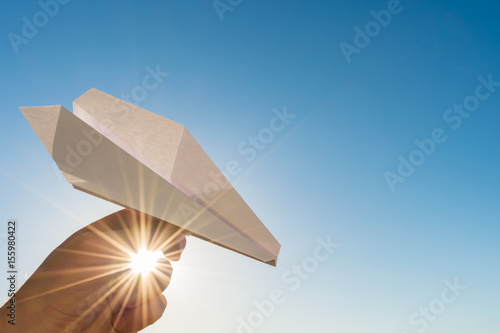 Hand holds white paper plane on blue sky and sun