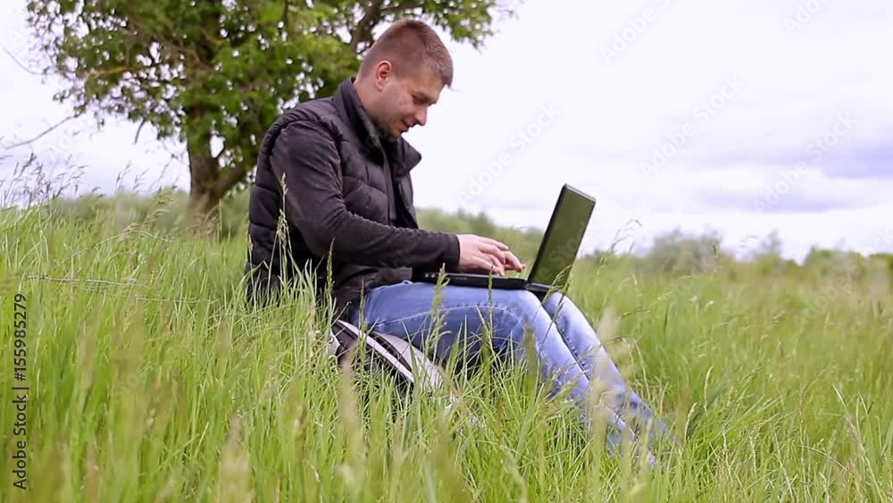 Young man sitting on the grass is working on laptop. Concept freelancer working sitting in the park. Young man sitting on the grass is working on laptop. Concept freelancer working sitting in the park.
