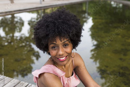 portrait of  a Happy young beautiful afro american woman sitting on wood floor and smiling. water background. Spring or summer season. Millennial. Casual clothing.
