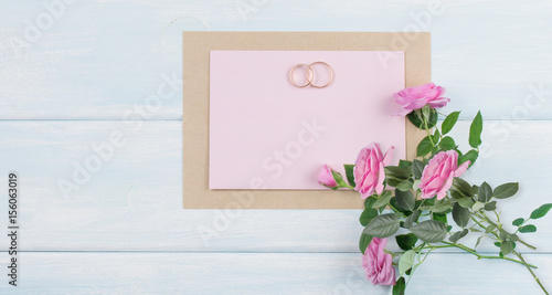 Pink roses and bridal rings with paper greeting card for wedding on background of shabby wooden planks.