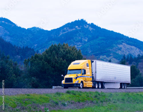 Cargo semi truck reefer trailer yellow on green highway winding between hill and mountain