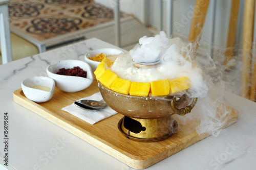 Canary Mango Bingsu is a popular Korean shaved ice dessert same as Kakigori, served in  pot with corn flakes cereal, red bean and sweetened condensed milk on the table and smoke of dry ice.