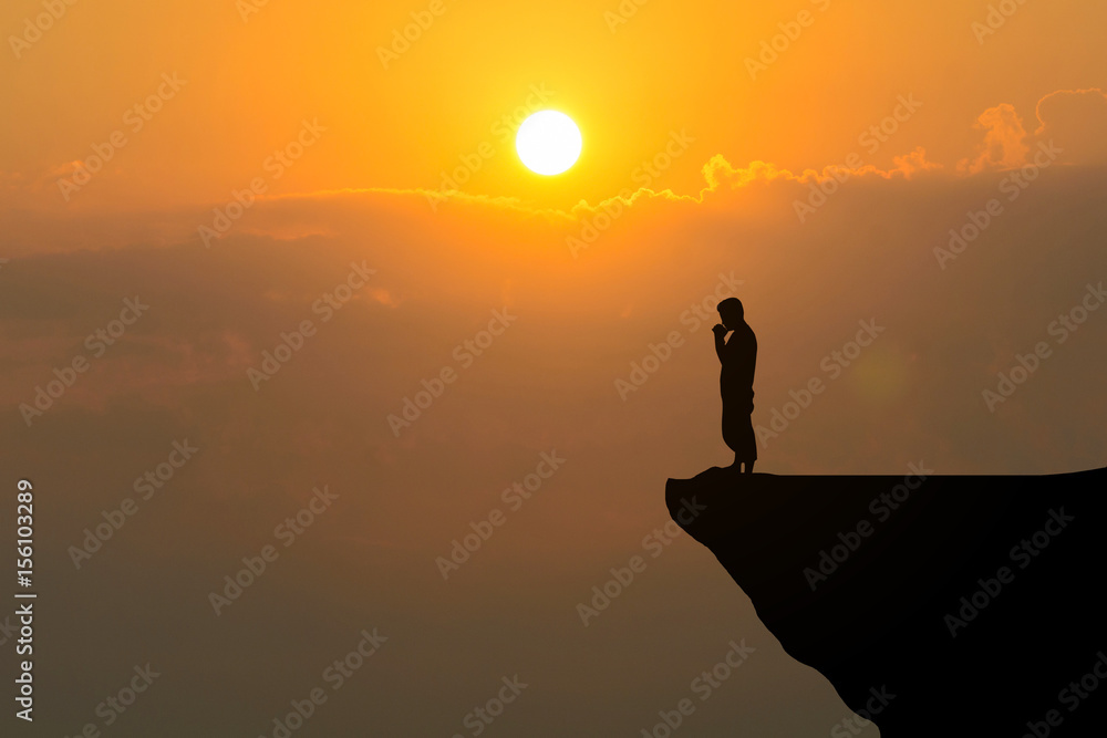 Man praying on cliff against sunset background