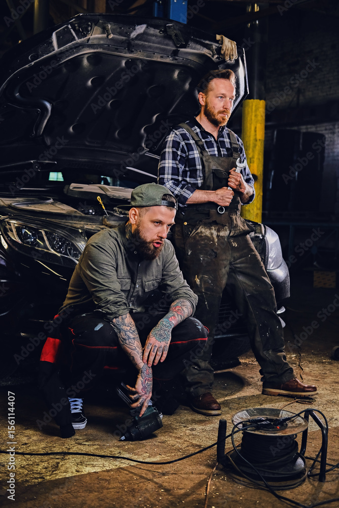 Two bearded tattooed mechanics near the car in a workshop. Two bearded tattooed mechanics near the car in a workshop.