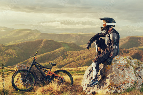 A professional downhill rider sits on a stone resting after a tense race on a mtb bike photo