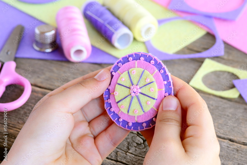 Fototapeta premium Small child made a beautiful flower from felt circles and beads. Child holds a felt flower in hands. Felt flower DIY, craft supplies on wooden table. Easy and fun sewing. Teaching kids to sew by hand