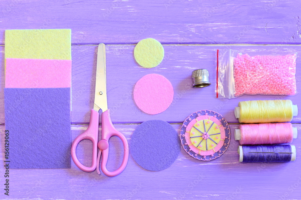 Fototapeta premium Pretty round flower made of felt and beads, colored felt circles and sheets, scissors, thread spools set, needle, pink beads on a wooden table. Sewing guide. Teaching sewing for children. Top view