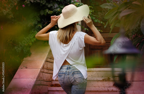 Beautiful blond woman in jeans, white tunic and big summer hat back view, fashion model portrait outdoors in the park with green leaves background and pink flowers, recreation walk in the garden