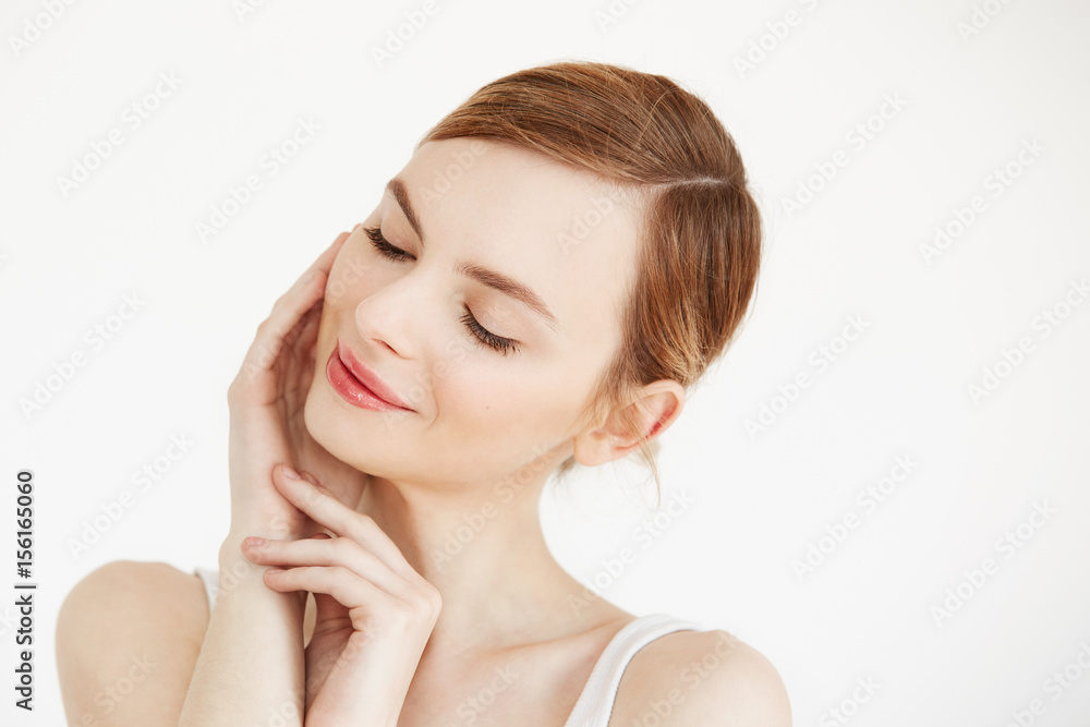 Portrait of young beautiful girl smiling with closed eyes touching face over white background. Facial treatment. Beauty cosmetology and skincare. Portrait of young beautiful girl smiling with closed eyes touching face over white background. Facial treatment. Beauty cosmetology and skincare.