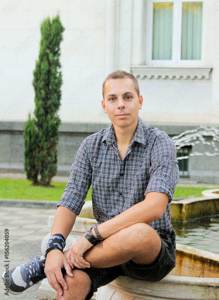 Obraz premium Man sitting on the stone fountain. Young man smile on a summer day in shirt