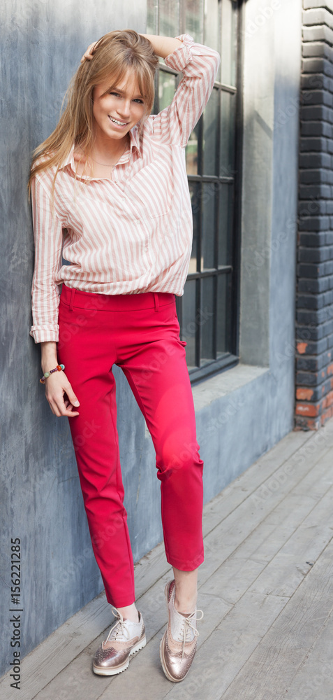 Portrait of a young woman dressed in a blouse, red Chino Trouser, and gold brogues. Posing next to a gray wall. Fashion. Portrait of a young woman dressed in a blouse, red Chino Trouser, and gold brogues. Posing next to a gray wall. Fashion.