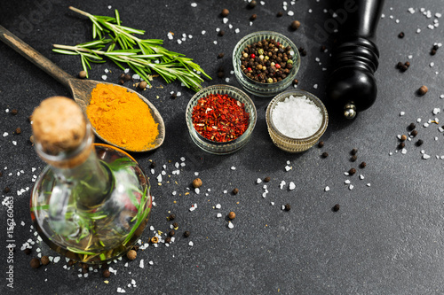 On a black stone surface of spices, black pepper mill, wooden spoons, in glass bowls pepper peas, salt, paprika, rosemary, Bottle with olive oil