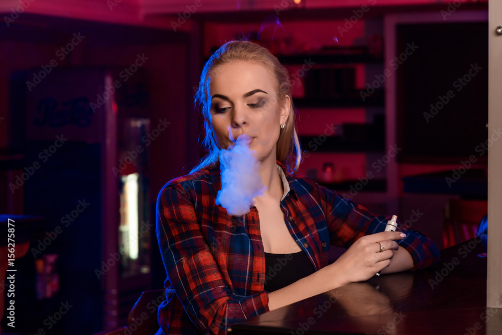 Young pretty woman in a shirt in a cage smoke an electronic cigarette at the vape bar Young pretty woman in a shirt in a cage smoke an electronic cigarette at the vape bar