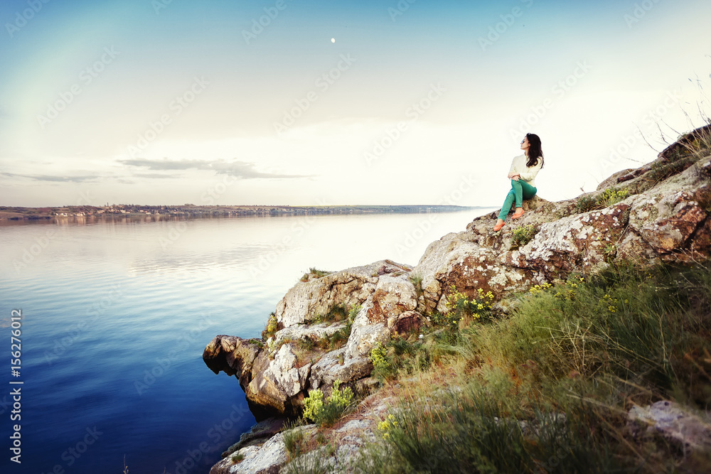 Young woman sits on rock and enjoys view of  river at sunset