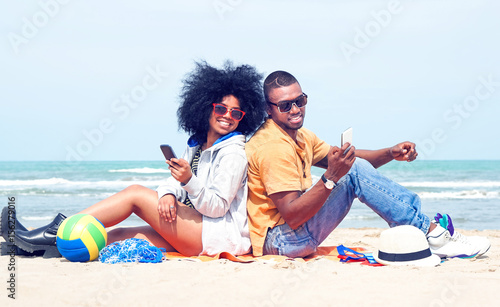 Happy afro american couple using mobile phone back to back on the beach  