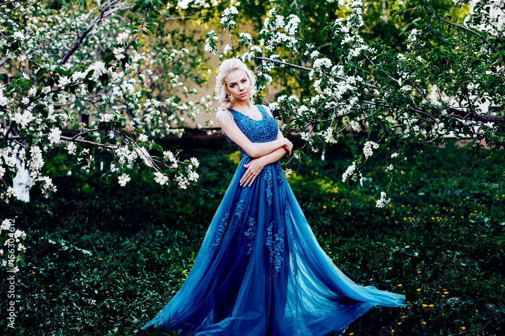 Beautiful young girl in a long evening blue dress in an apple blossoming garden
