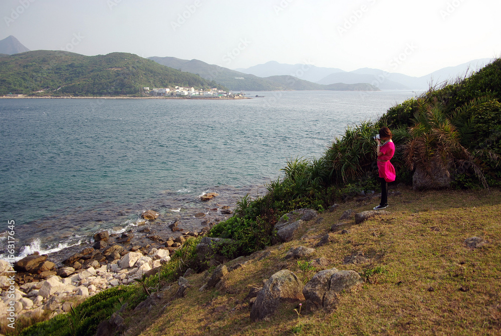 A girl in reddish pinkish dress on the cliff taking picture of the sea