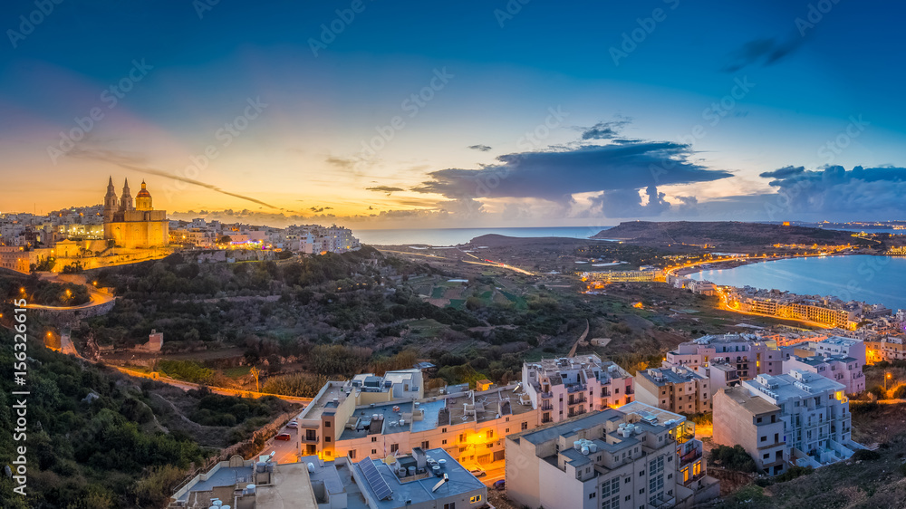 Naklejka premium Il-Mellieha, Malta - Beautiful panoramic skyline view of Mellieha town at blue hour with Paris Church and Mellieha beach and Gozo at background with blue sky and clouds