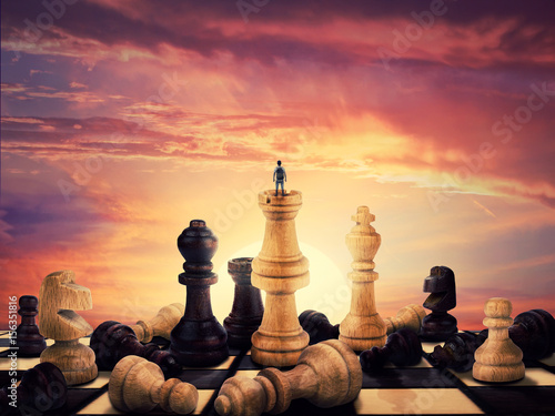 Successful boy standing on a chess piece meeting the sunrise on the horizon. Gigant chess figures scattered on the vintage chessboard. Symbol of business aspirations, freedom and leadership concept. 