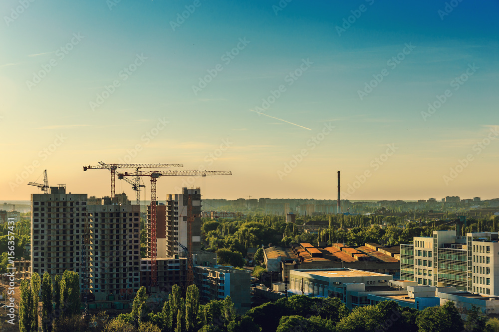 Fototapeta premium Urban scene with construction industry, cranes, building, houses, city skyline in sunset,