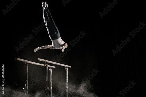 Male athlete performing difficult exercise on gymnastic parallel bars with talcum powder. Isolated on black.