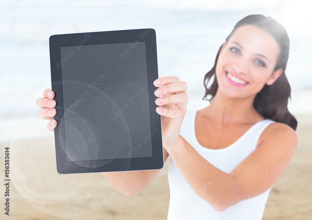 Smiling woman holding out tablet against blurry beach with flare